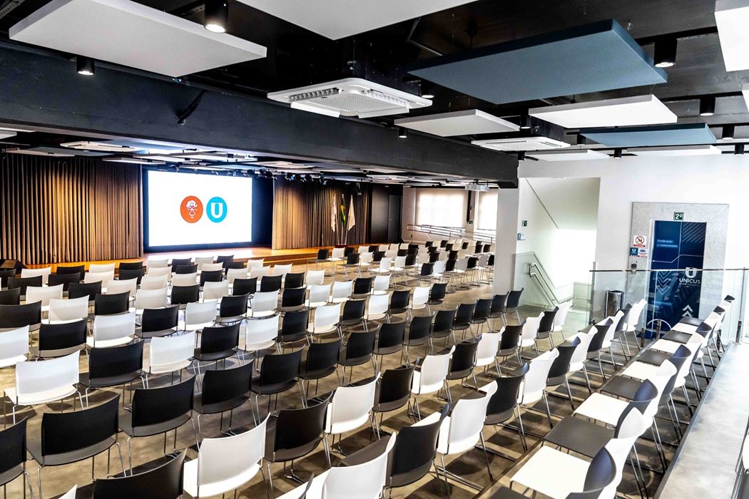 Auditório do grupo, com poltronas em fileiras e a marca das duas escolas projetada no telão