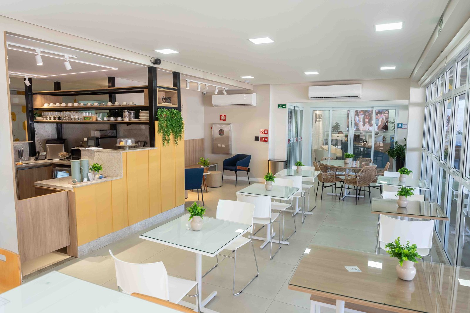 Café e coworking das famílias, com balcão amarelo, mesas de vidro e vista para a área envidraçada