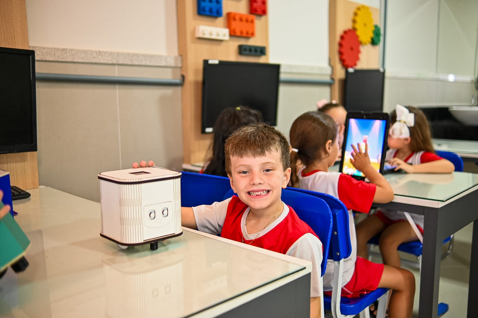 Menino sorrindo ao lado de um robô de mesa na sala de tecnologia, com colegas usando tablets ao fundo