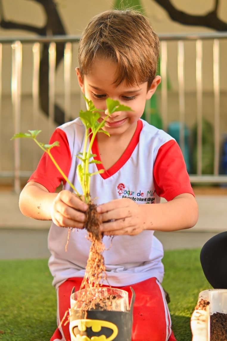 Menino de uniforme do Toque de Mãe plantando uma muda em um vaso feito de garrafa reaproveitada