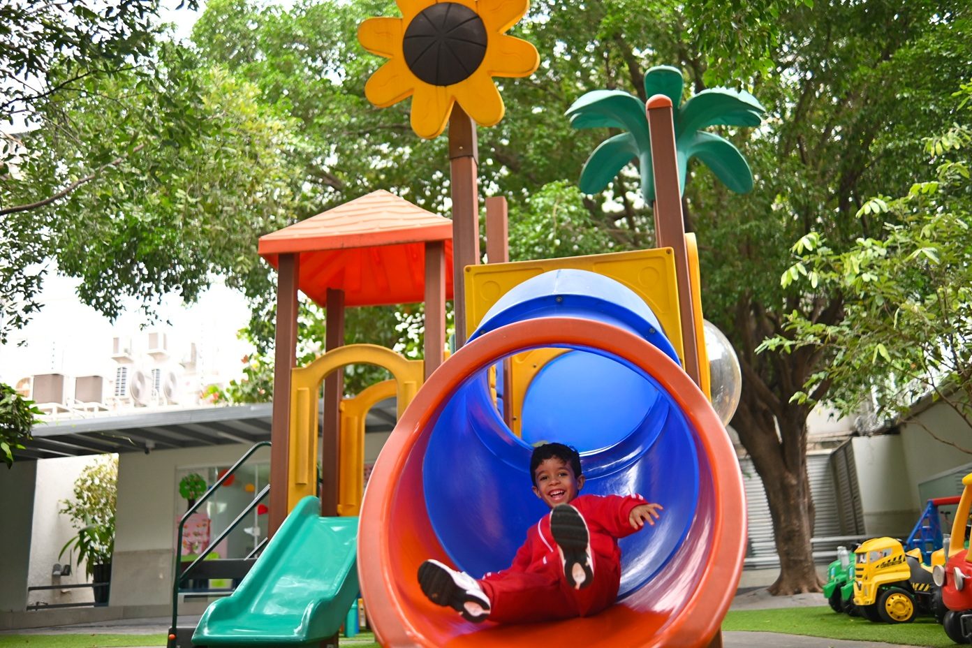 Criança descendo o tubo colorido do playground, entre árvores e brinquedos de montar