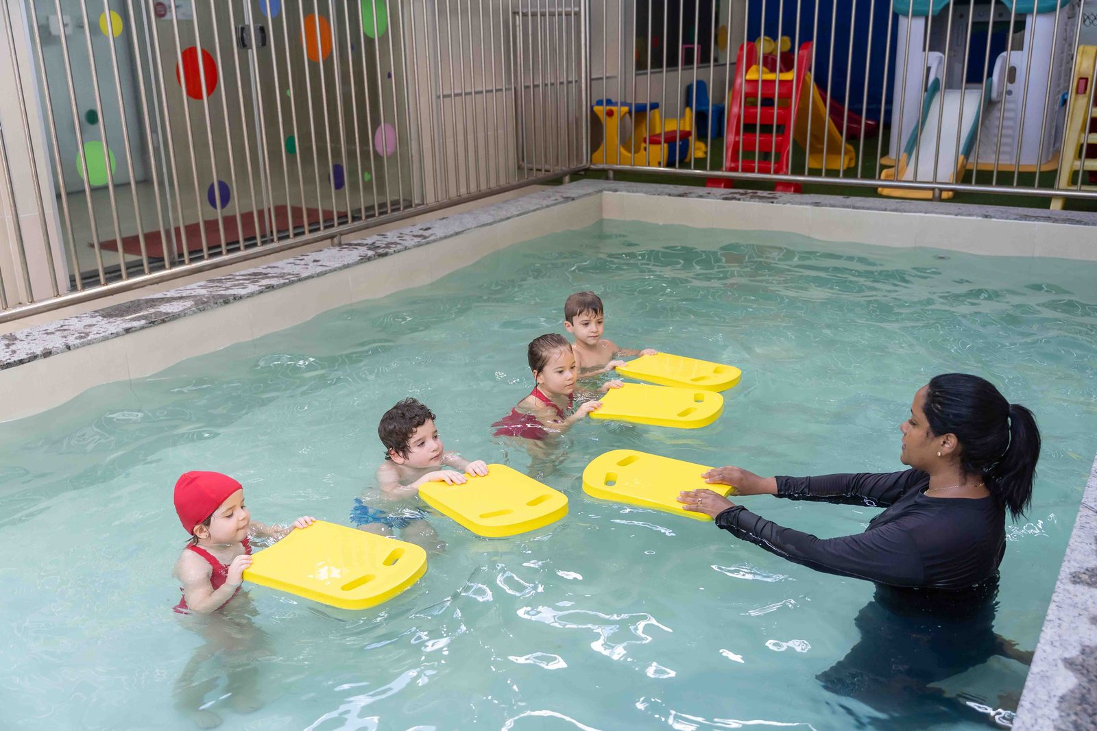 Aula de natação na piscina aquecida, com quatro crianças e a professora usando pranchas