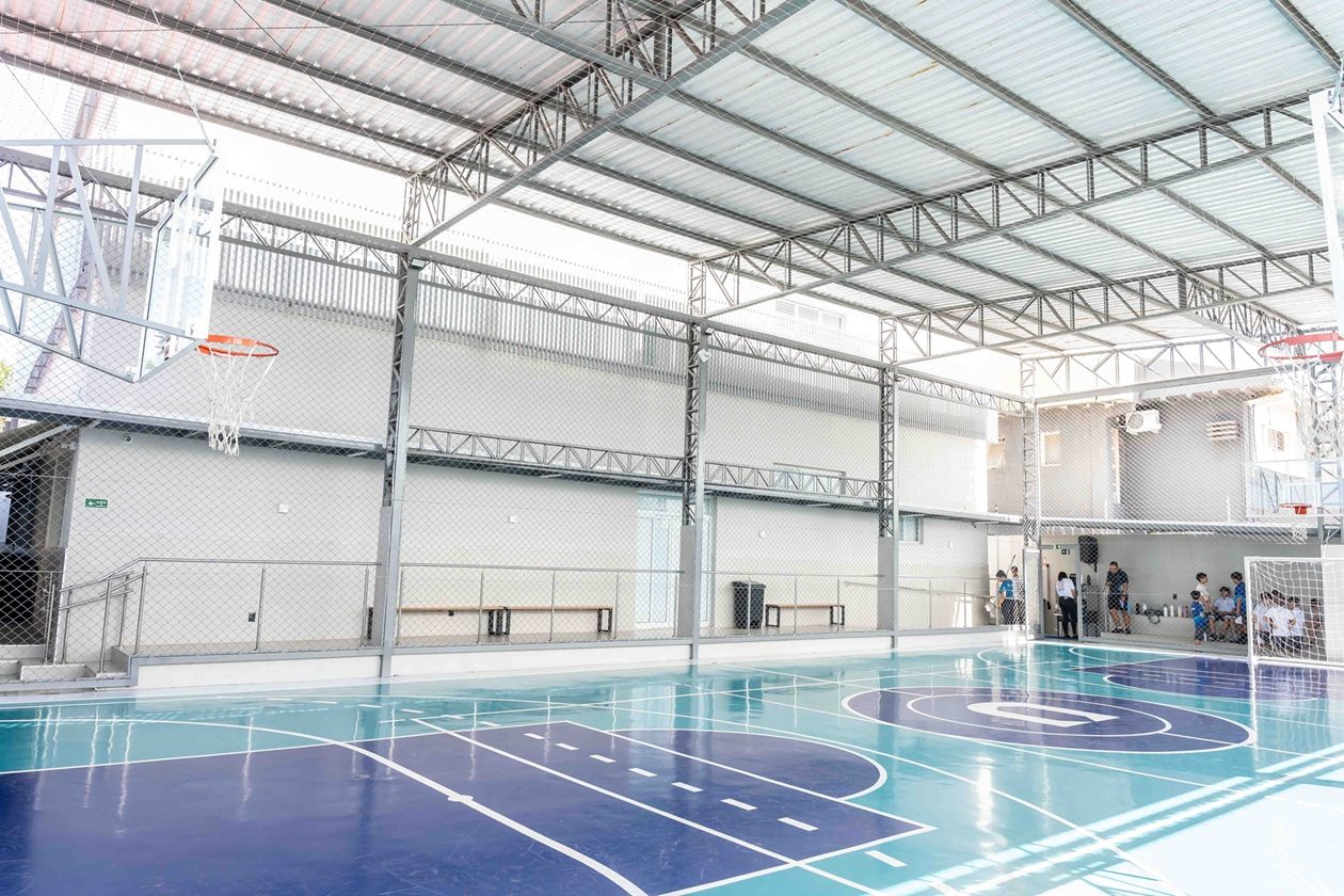 Quadra de esportes coberta, com piso azul e turquesa, tabelas de basquete e traves de futsal