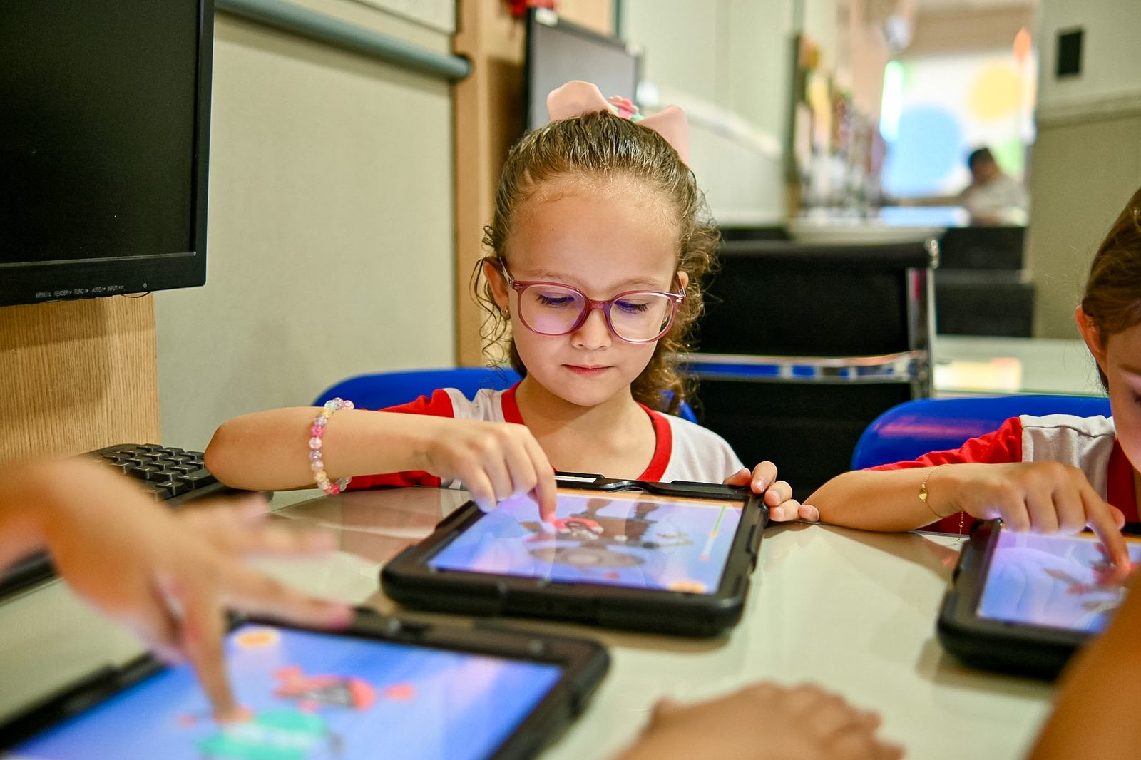 Menina de óculos usando um tablet na mesa da sala de tecnologia, ao lado de colegas