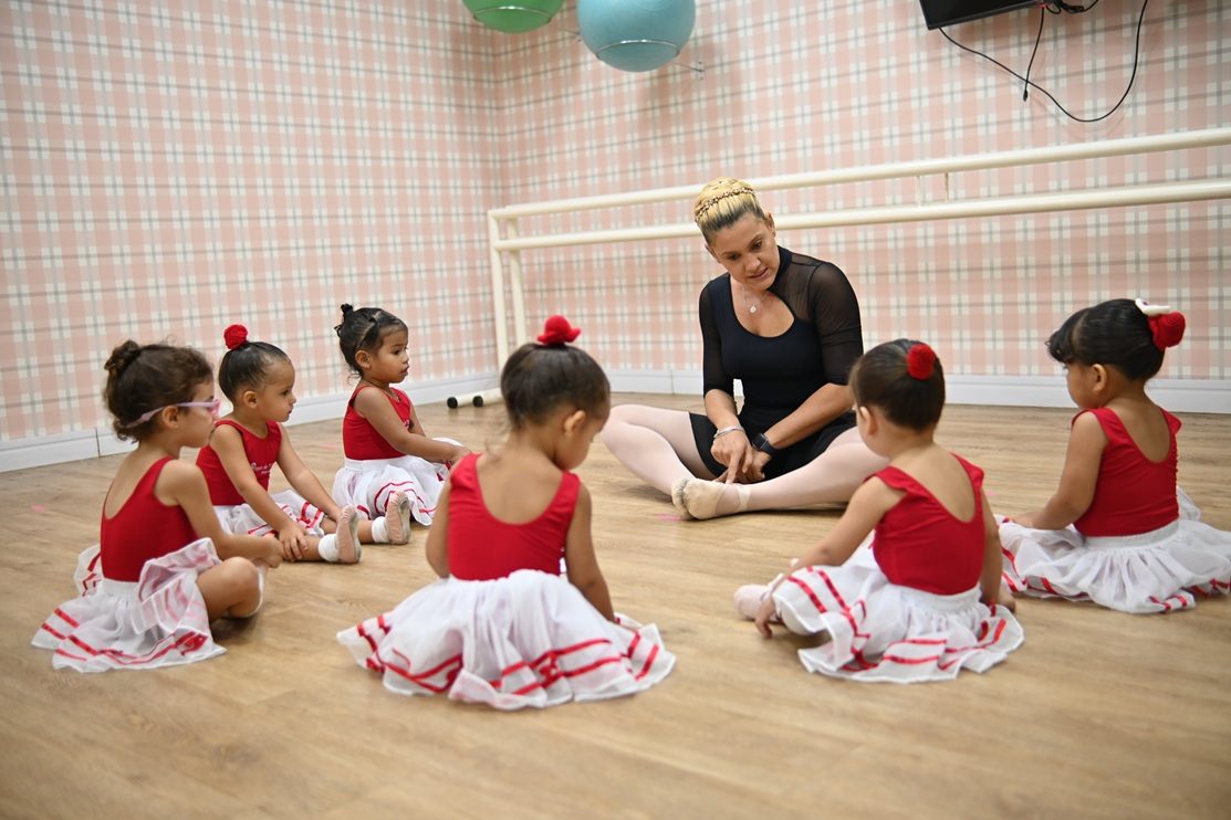Aula de ballet: seis crianças de tutu sentadas em roda no piso de madeira, com a professora