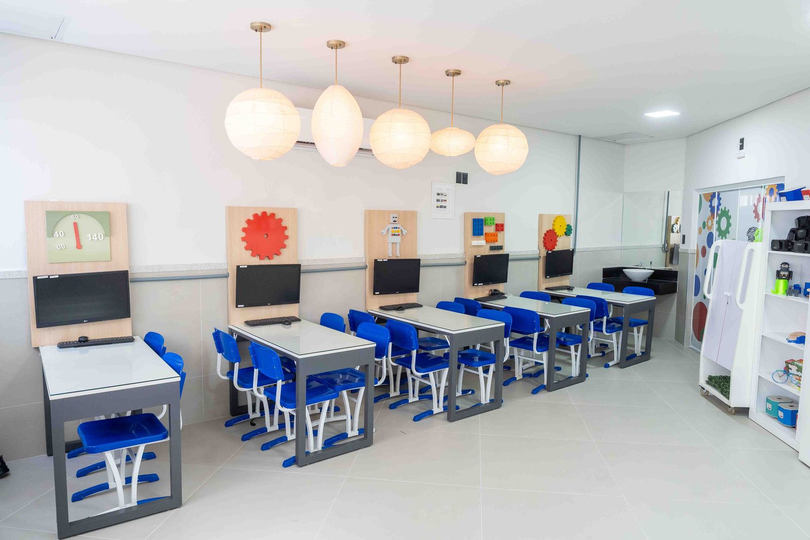 Sala de robótica com bancadas, computadores e painéis de engrenagens e peças de montar nas paredes